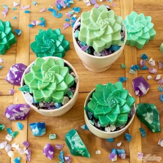 Three green succulent-shaped soaps in white pots sit on a wooden surface, surrounded by colorful gemstones in purple and blue, creating a vibrant, whimsical tone.