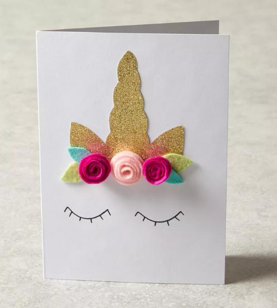 A handmade greeting card features a glittery golden unicorn horn, felt flowers, and closed eyes. The card conveys a whimsical, magical tone.