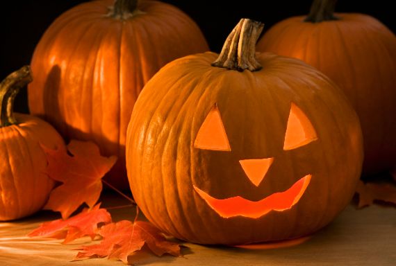 Origin-of-Jack-O’-Lanterns-Halloween-2 Three pumpkins on a table, one carved into a glowing jack-o'-lantern with triangular eyes and a wide smile. Autumn leaves scattered around evoke a festive Halloween atmosphere.
