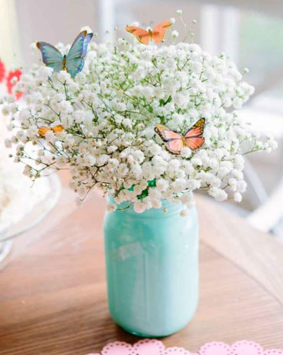 A bouquet of delicate white baby's breath flowers sits in a light blue jar on a wooden table, adorned with colorful butterfly decorations, evoking a cheerful spring vibe.