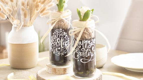 Two glass Easter-decor_Upcycled-vase-with-lettering with budding flowers and Easter greetings, tied with ribbons, sit on wooden coasters. Soft sunlight and dried flowers create a warm, festive ambiance.