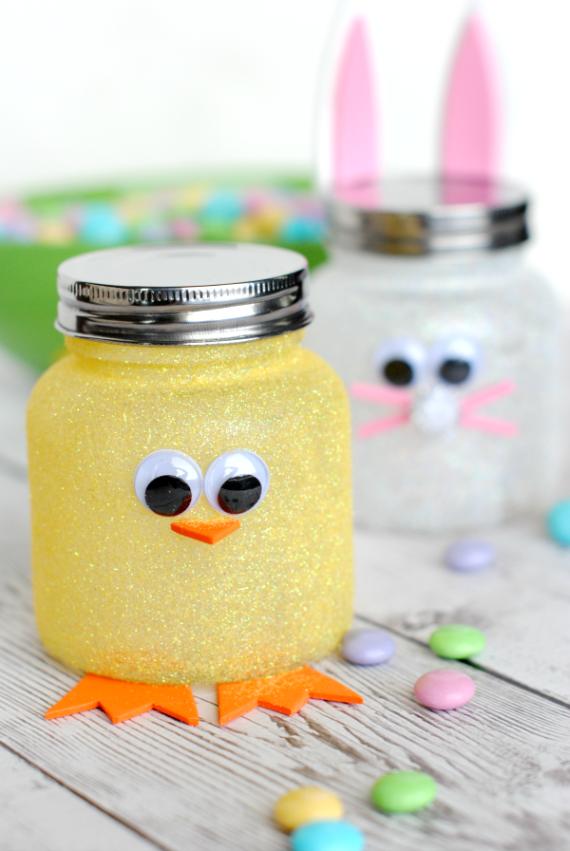 Glittery yellow jar decorated as a chick with googly eyes and orange beak and feet. Behind it, a white jar with bunny ears. Pastel candies scattered nearby.