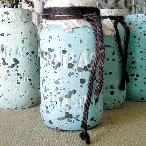Light blue Mason jars with gray paint splatters are wrapped with dark yarn near the top. They are set against a fabric background, conveying a rustic, artistic vibe.