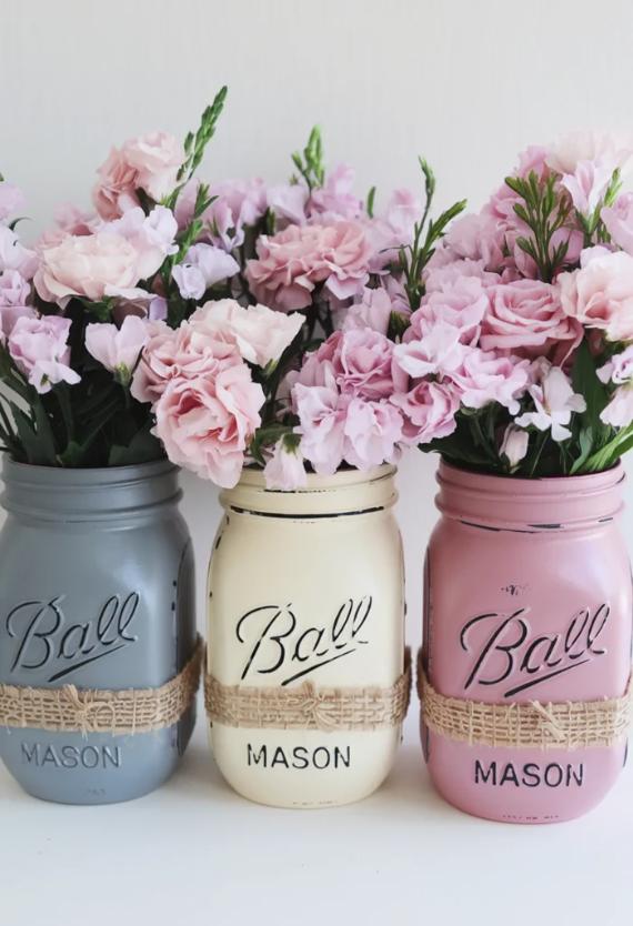 Three Mason jars in gray, cream, and pink hold bunches of light pink flowers, accented with greenery. The jars are wrapped with burlap, creating a rustic, charming look.