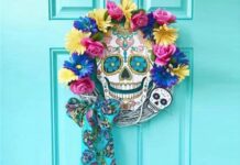 Halloween Door Decoration Ideas A white grapevine wreath decorated with bright pink, yellow, and blue flowers, a central sugar skull sign, and a matching turquoise skull ribbon on a turquoise door.