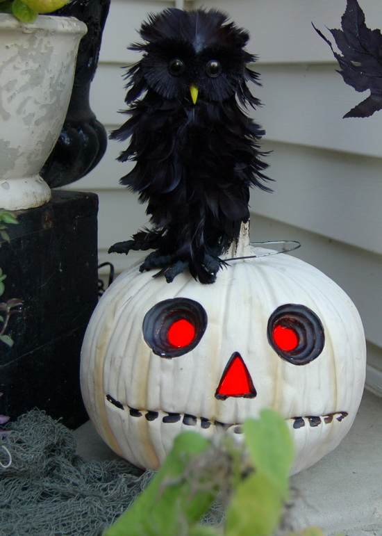 A fluffy black faux owl prop perched on top of a carved white pumpkin jack-o'-lantern with glowing red eyes and nose.