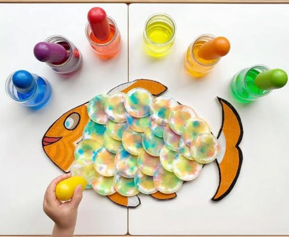 Child assembling colorful fish scales from painted cotton pads, surrounded by jars of vibrant paint with droppers, creating a playful, creative scene.