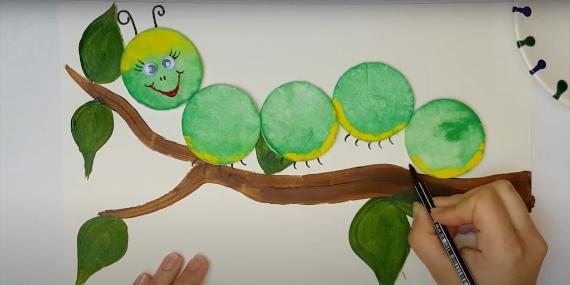 A hand is drawing a green Cotton Pad Caterpillar on a branch with leaves. The caterpillar has a smiling face and is made of green circles. The scene is playful and creative.