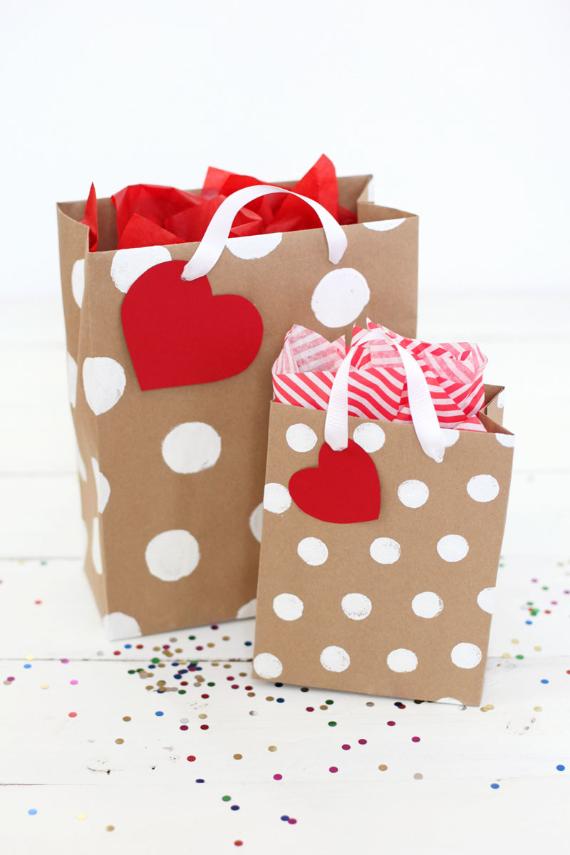 Two brown paper gift bags with white polka dots and red heart tags. They contain red and striped tissue paper, with colorful confetti on a white surface.