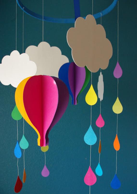 A colorful paper mobile hangs against a teal wall, featuring clouds, a hot air balloon, and raindrops in vibrant hues, creating a whimsical and playful tone.