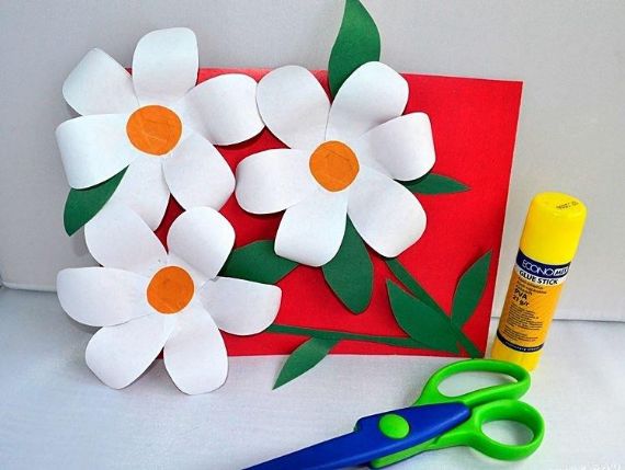 A craft project with three paper daisies on a red card, accompanied by a yellow glue stick and green scissors