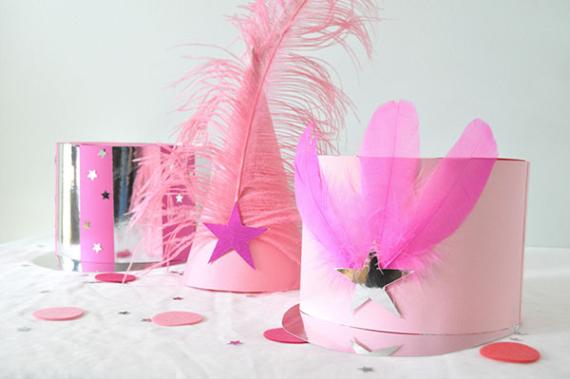 Three whimsical hats on a white surface: a pink cone with feathers and a purple star, a pink feathered hat, and a glittery silver top hat with stars. Playful and festive.