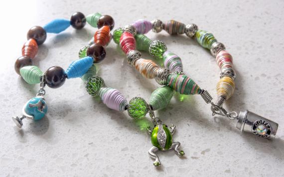 Three creative paper craft ideas for holidays and celebrations of colorful beaded bracelets with unique charms, including a globe, frog, and cup, lie on a light surface, conveying a playful and vibrant mood.
