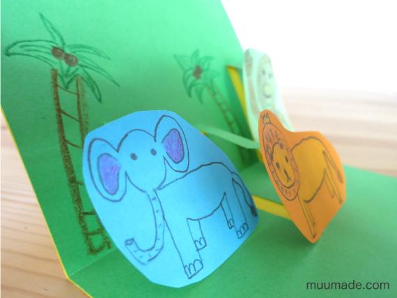 Pop-up card with colorful paper animals, including a blue elephant and an orange lion, set against a green background with drawn palm trees.