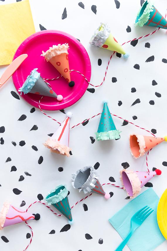 A festive, playful scene with colorful party hat garland on a speckled table. Includes a pink plate, pastel napkins, and utensils, conveying a cheerful vibe.