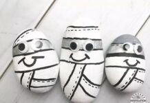 35 Simple Halloween Crafts For Kids on the scariest night Three painted rocks resembling cartoon mummies, with black and white colors, displaying googly eyes and smiling faces, arranged on a light wooden surface.