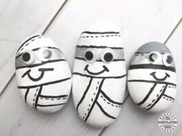 Three painted rocks resembling cartoon mummies, with black and white colors, displaying googly eyes and smiling faces, arranged on a light wooden surface.