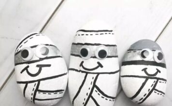 35 Simple Halloween Crafts For Kids on the scariest night Three painted rocks resembling cartoon mummies, with black and white colors, displaying googly eyes and smiling faces, arranged on a light wooden surface.