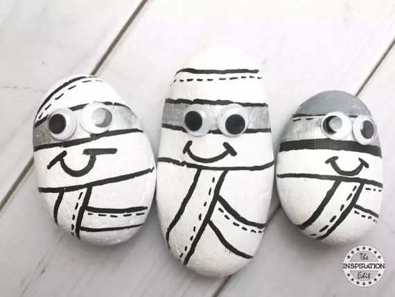 Stone-Crafts-for-Halloween Three painted rocks resembling cartoon mummies, with black and white colors, displaying googly eyes and smiling faces, arranged on a light wooden surface.