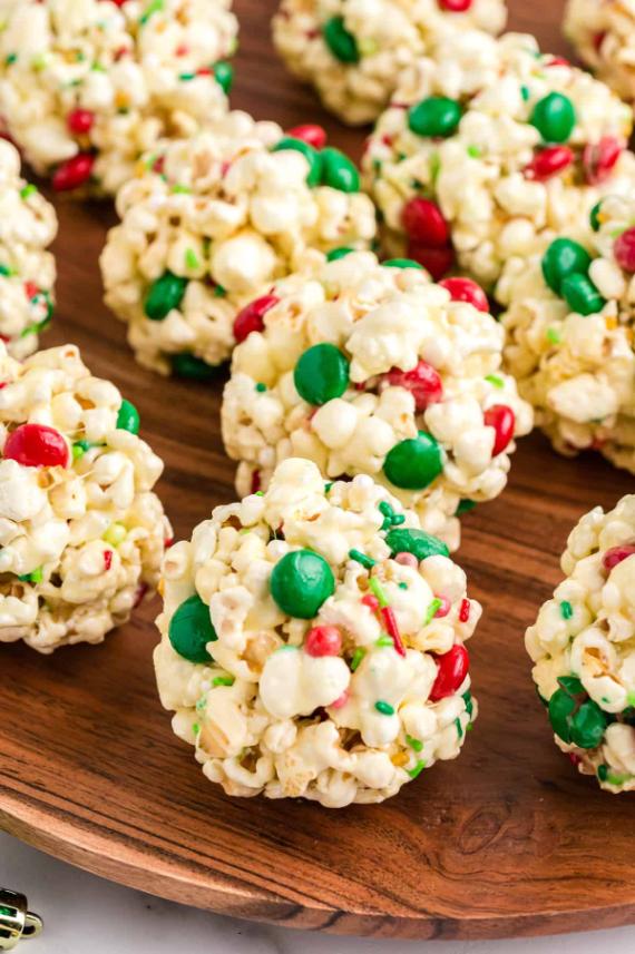 Colorful popcorn balls coated in white chocolate, decorated with red and green candies, served on a wooden platter.