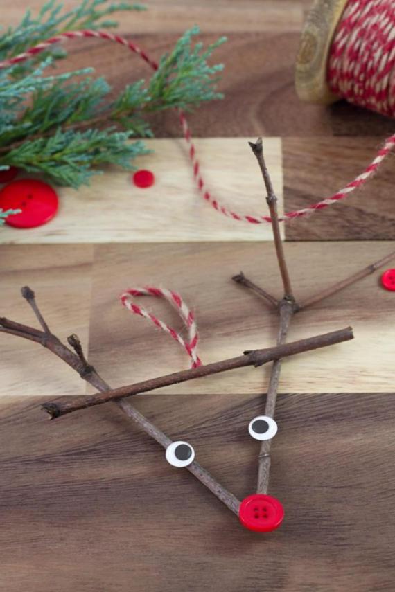 A whimsical reindeer face created from twigs, buttons, and string, set on a wooden surface with greenery in the background.