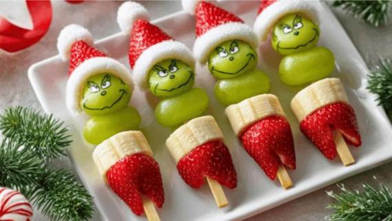 Holiday-themed fruit skewers featuring green grapes and strawberries topped with Santa hat decorations and Grinch faces.
