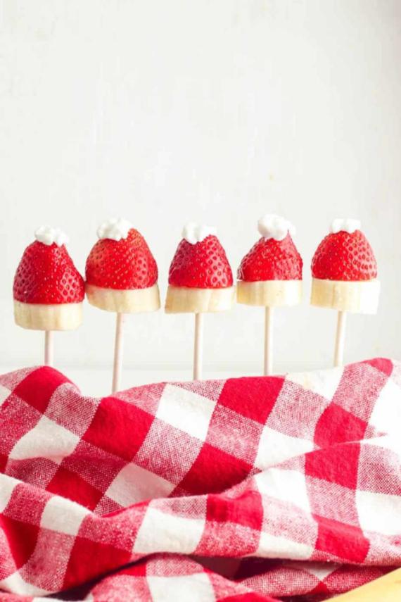 Strawberries with whipped cream hats on banana slices, arranged on sticks, set against a red and white checkered cloth.