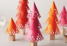 17 0f Our Favorite Paper Crafts for Adults Colorful, paper-cut cone trees stand on wooden bases, resembling a forest. The hues are pink and orange, surrounded by small decorative deer figures.