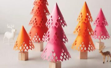 17 0f Our Favorite Paper Crafts for Adults Colorful, paper-cut cone trees stand on wooden bases, resembling a forest. The hues are pink and orange, surrounded by small decorative deer figures.