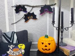Three handmade black felt bats with colorful neon borders hanging from a natural wooden branch on a textured gray wall.