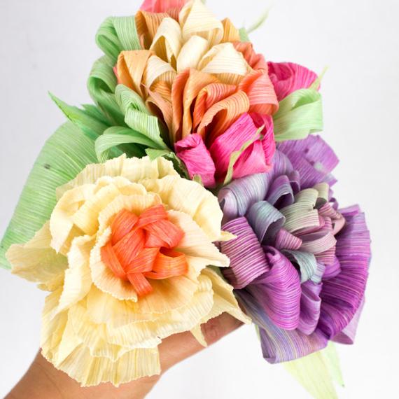 A hand holds a vibrant bouquet of multicolored, textured paper flowers. The arrangement features soft hues of yellow, orange, pink, purple, and green.