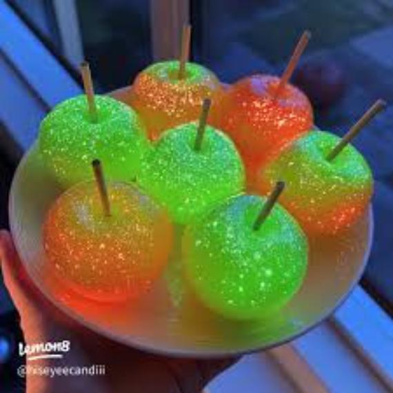 Glowing candy apples in neon green and orange on a plate, each with a stick, against a dark background. Theses Fall decor with unexpected colors are vibrant and festive.