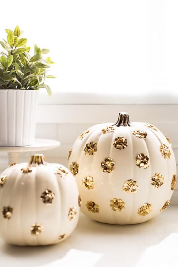White pumpkins adorned with gold flower motifs sit beside a potted plant on a sunlit windowsill, creating an elegant Jewel-toned fall decorating ideas and festive autumnal display.