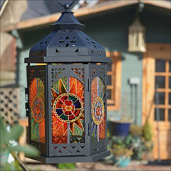 A decorative lantern with intricate stained glass panels in vibrant orange and green hues hangs outdoors. A blurred wooden house is in the background.