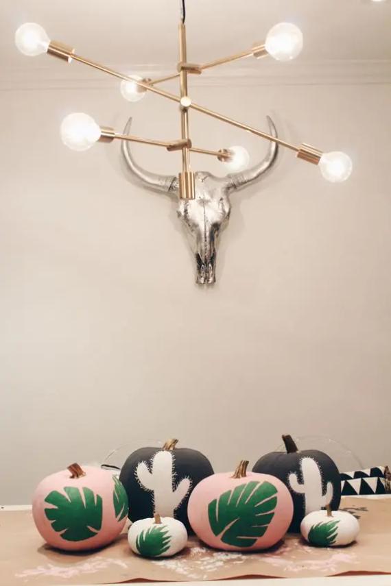 A modern room with a brass chandelier and silver animal skull on the wall. Below, decorated with Pastel pumpkins decor with green leaf and cactus designs sit on a table.