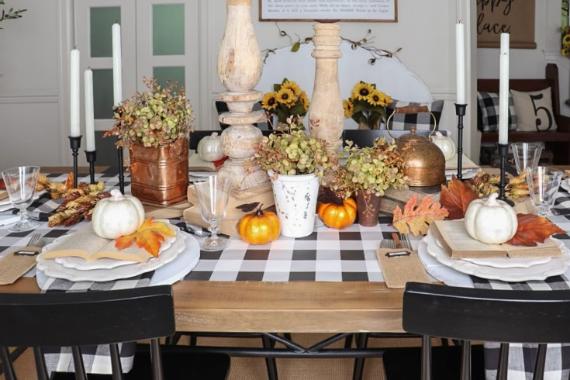 A fall-themed dining table with checkered cloth, small white pumpkins, dried flowers, candles, and autumn leaves evokes a cozy, festive atmosphere.
