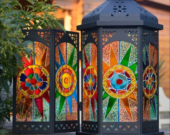 Colorful stained glass lantern with intricate geometric patterns and vibrant colors stands outdoors, casting a warm, inviting glow in a garden setting.