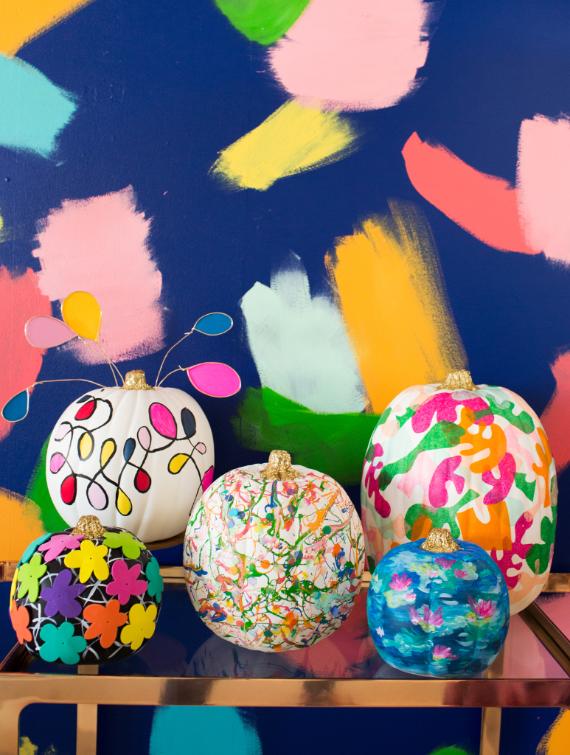 Colorful Fall decor with unexpected colors, hand-painted pumpkins in abstract designs sit on a reflective surface, against a vibrant multicolored background with bold brushstrokes.