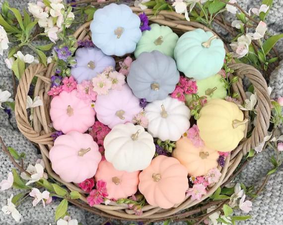 A basket filled with Pastel pumpkins decor surrounded by vibrant pink and purple flowers, set on a soft gray knitted fabric and leafy twigs.
