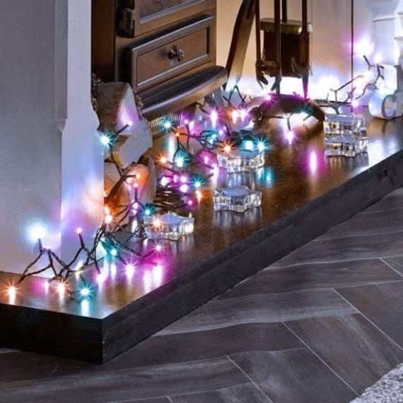 A cozy fireplace setting with colorful string lights in blue, pink, and orange hues. Glass candleholders scatter warm reflections, creating a festive ambiance.