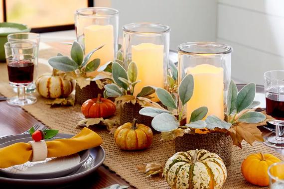 Thanksgiving table centerpiece with glowing candles in glass holders, surrounded by leaves and mini pumpkins. Warm, cozy autumn ambiance.
