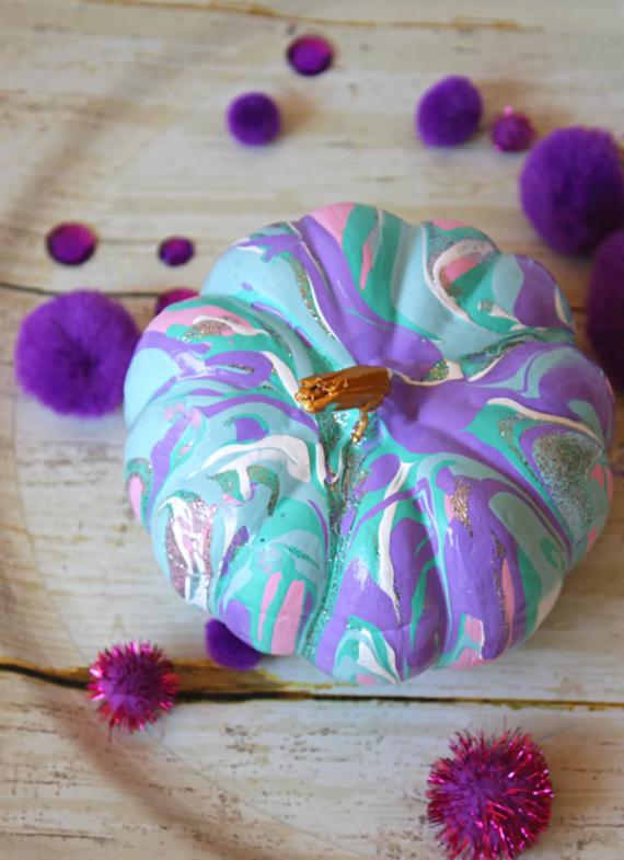 A marbled Pastel pumpkins decor in purple, and pink hues with a gold stem is surrounded by vibrant purple pom-poms on a rustic wooden surface.