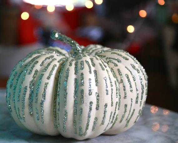 White pumpkin adorned with vertical silver glitter stripes on a marble surface. Soft, warm bokeh lights in the blurred background convey a festive mood.