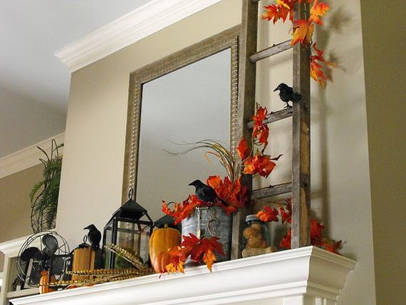 Autumn-themed mantel decor featuring vibrant orange leaves, pumpkins, black crows, and a rustic ladder. A mirror reflects light, adding warmth.