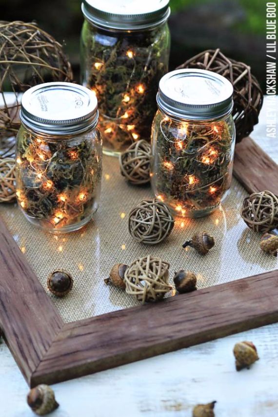 Three mason jars filled with moss and twinkling fairy lights sit on a wooden tray. Surrounding the jars are woven wicker balls and scattered acorns, creating a rustic, cozy atmosphere.