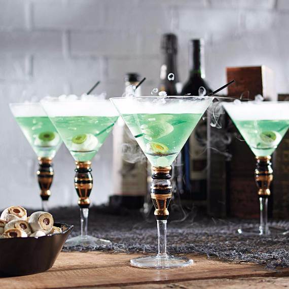 Four martini glasses with green cocktails, garnished with olives, emit a smoky effect. Background includes blurred bottles and a wooden surface.