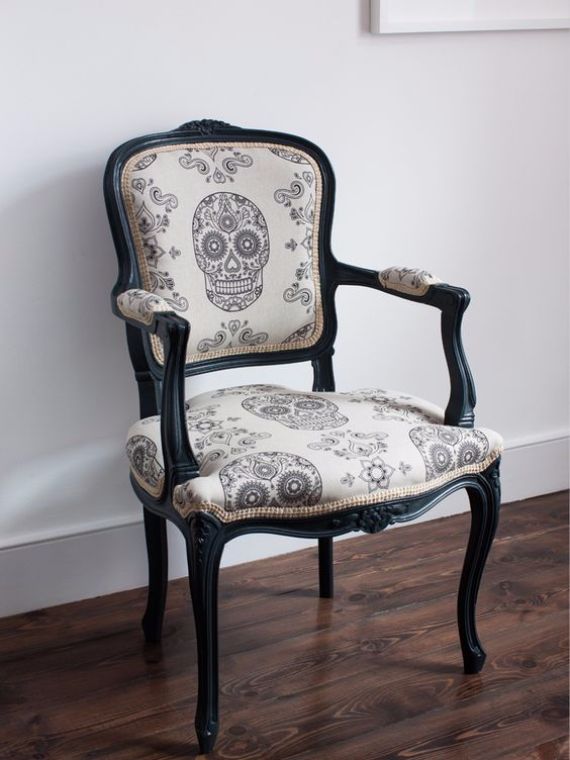 Vintage wooden chair with black frame, featuring beige upholstery with intricate skull designs. It sits on a wooden floor, creating a gothic yet elegant tone.