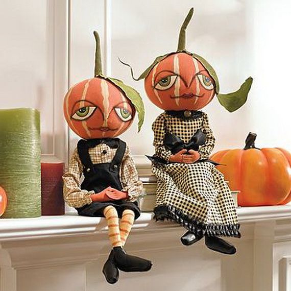 Two whimsical pumpkin-head dolls sit on a mantel. Dressed in vintage attire, they evoke a playful autumn vibe, surrounded by colorful candles and a pumpkin.