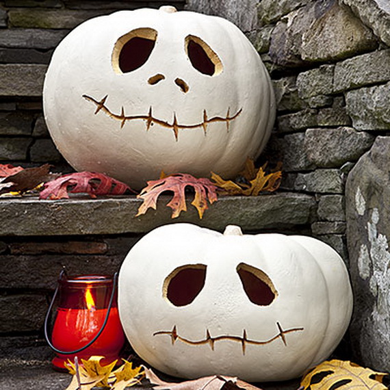 Two white pumpkins carved with playful, stitched smiles and large eyes sit on stone steps with scattered autumn leaves and a glowing red candle.