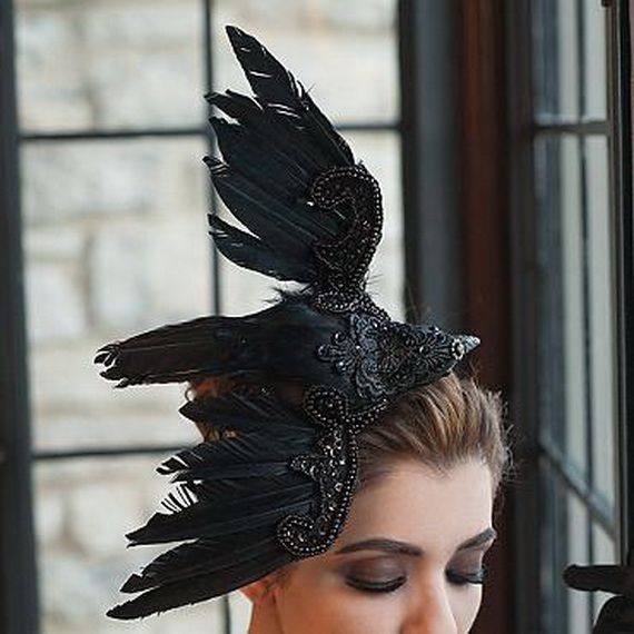 A woman wearing an elaborate black fascinator adorned with feathers and beads that form a bird shape. The background shows a window with stone walls.